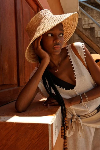 A young Black woman poses in natural light, wearing a straw sunhat and light-colored linen outfit, leaning against a wood structure.