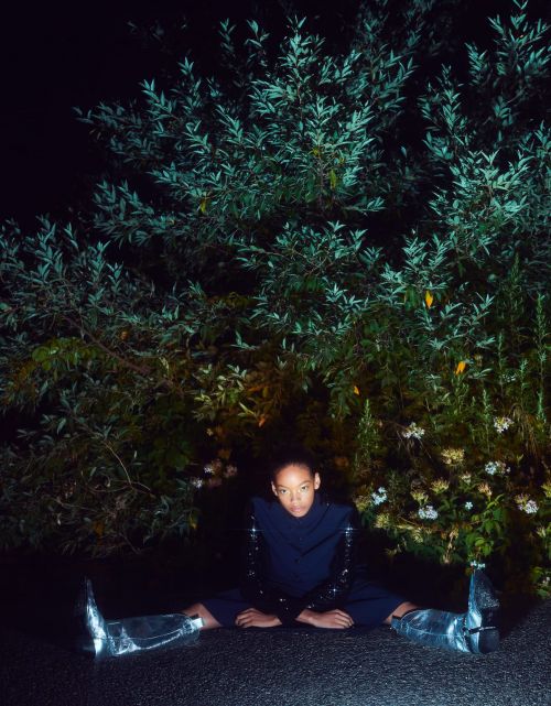 Woman with afro sits on ground with spread legs in blue suit, sequin sleeves, and silver boots in front of large green bush.