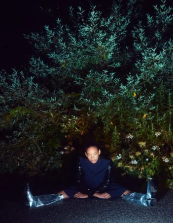 Woman with afro sits on ground with spread legs in blue suit, sequin sleeves, and silver boots in front of large green bush.