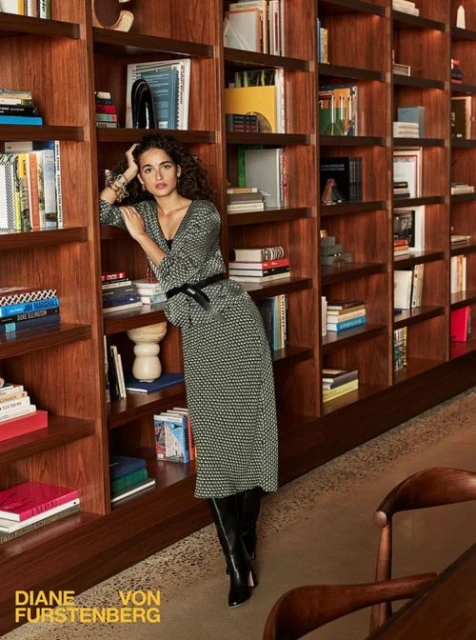 Adult female posing in front of a bookshelf, wearing a black-and-white dress and black boots, in an indoor setting.