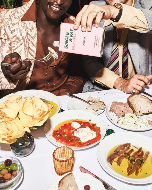 Two men at a table pouring organic olive oil on a pasta dish, surrounded by various plates of food, including tomatoes and anchovies.