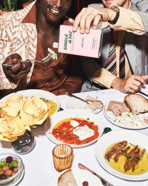 Two adult male models at a dinner table pour olive oil over pasta, featuring dishes of food and a pink olive oil bottle.