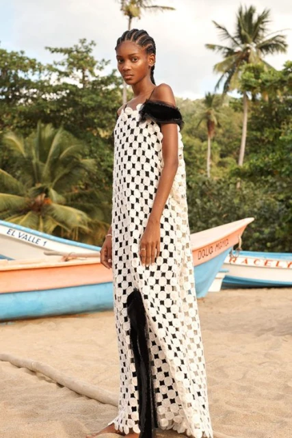 A slender, dark-skinned woman with braided hair is the main focus, wearing a white, patterned maxi dress, standing on a beach near boats.