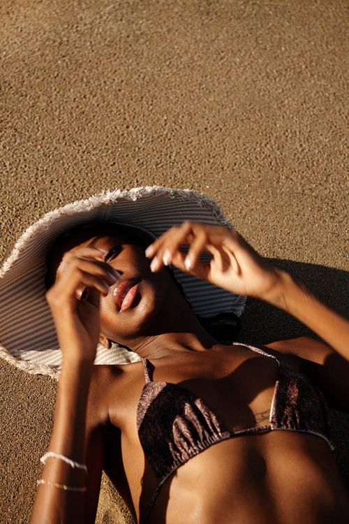 Woman in bikini and hat lying on tan sand, arms raised toward the sun. Relaxing on the beach on a sunny day. Fashion, summer, travel.