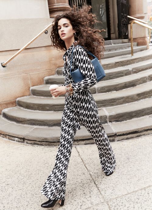 A woman in patterned shirt and pants strides on city sidewalk with a blue purse. Steps and building facade in the background.