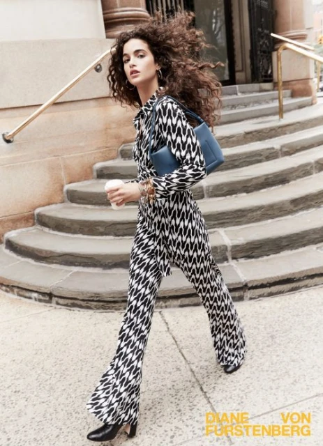 A young woman in patterned wide-leg pants walks outdoors, carrying a blue purse and coffee, with city buildings in the background.