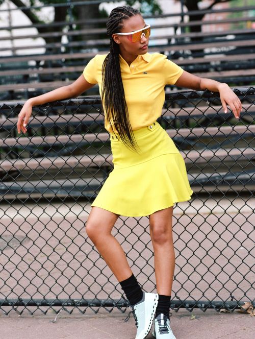A woman with braids in a yellow top and skirt leans against a fence wearing sunglasses and white sneakers, posing outdoors in daytime.