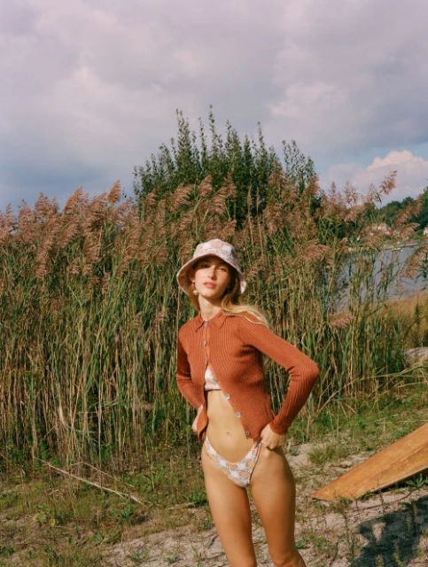 Woman poses in bikini and cardigan by tall reeds in a natural setting under a cloudy sky, wearing a floral bucket hat.