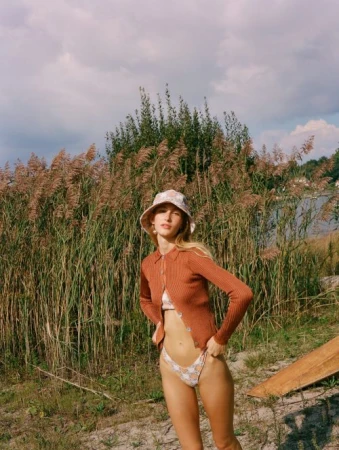 Woman poses in bikini and cardigan by tall reeds in a natural setting under a cloudy sky, wearing a floral bucket hat.