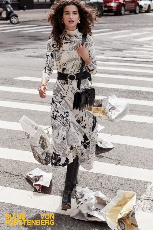 A stylish woman in a newspaper-print dress confidently strides across a crosswalk in an urban setting, surrounded by scattered papers.