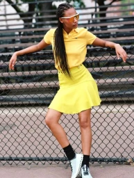 Model in yellow polo shirt and matching skirt leaning against a wire fence wearing futuristic sunglasses