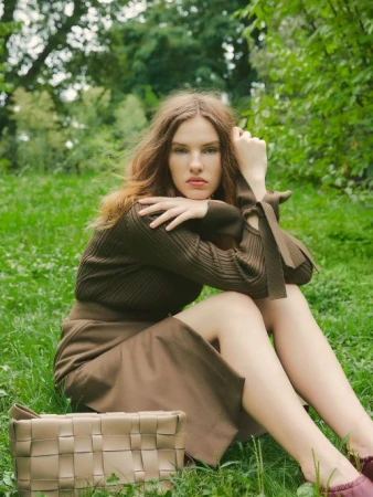 Woman in an olive green sweater and skirt seated on grass with a beige woven handbag