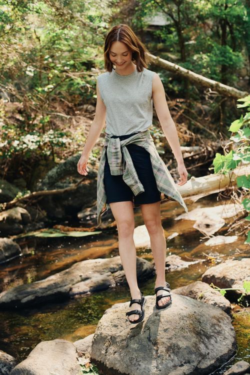 A woman in a gray tank top and black shorts stands on rocks in a stream with a plaid shirt tied around her waist in a forest setting.