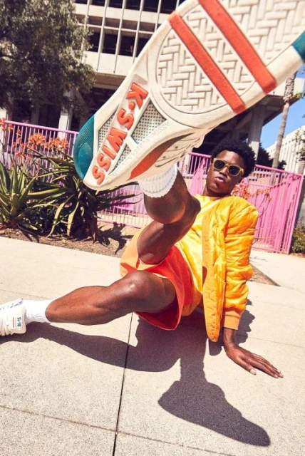 Person posing with a K-Swiss shoe, wearing an orange outfit and sunglasses, in an urban setting with buildings and trees.