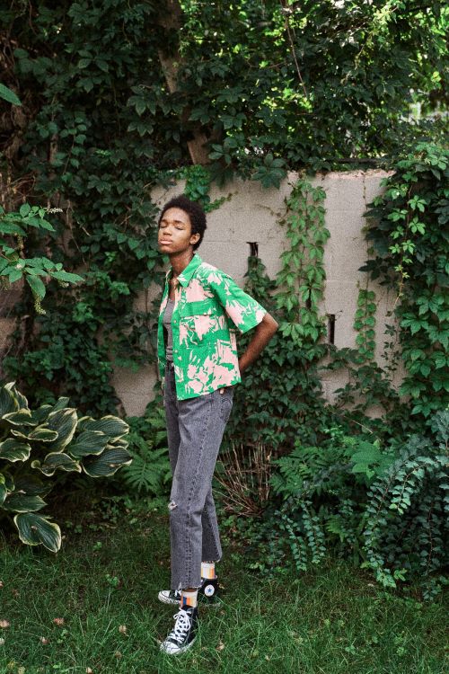 Woman in tropical-print shirt and black jeans poses in a green outdoor setting with natural light and black high-top sneakers.