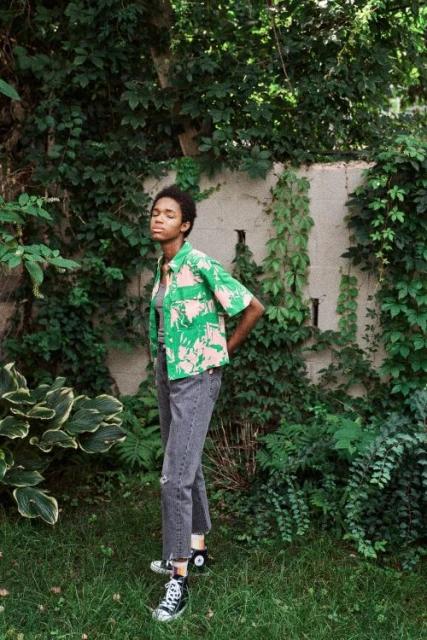 Woman in tropical-print shirt and black jeans poses in a green outdoor setting with natural light and black high-top sneakers.