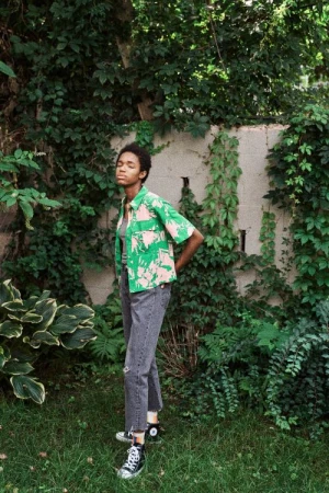 Woman in tropical-print shirt and black jeans poses in a green outdoor setting with natural light and black high-top sneakers.