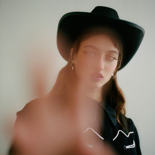 Portrait of a young adult woman wearing a black cowboy hat and black collared shirt, with hand blurred in foreground against white backdrop.