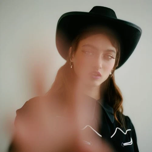 Portrait of a young adult woman wearing a black cowboy hat and black collared shirt, with hand blurred in foreground against white backdrop.