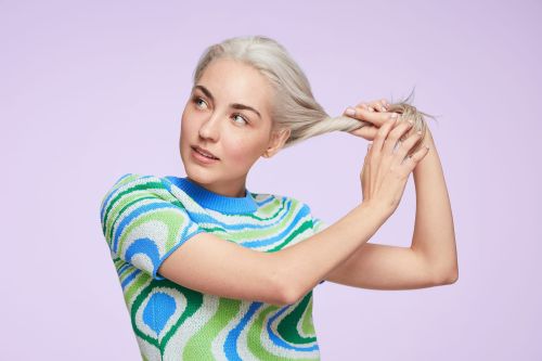 A young woman with platinum blonde hair is holding her hair and glancing to the left against a light purple backdrop in a studio shot.