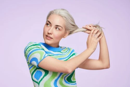 A young woman with platinum blonde hair is holding her hair and glancing to the left against a light purple backdrop in a studio shot.