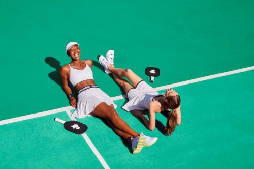Two female athletes relax on a vibrant green court in white athletic wear. Pickleball paddles rest nearby, captured in a bright, high-angle shot.