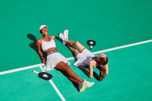 Two female athletes relax on a vibrant green court in white athletic wear. Pickleball paddles rest nearby, captured in a bright, high-angle shot.