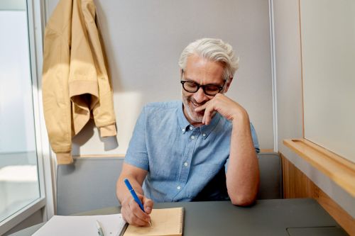 Grey-haired man wearing glasses seated at a desk indoors writing in a notebook with a pen and wearing a light blue shirt.