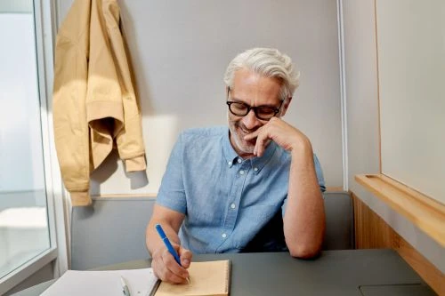 Grey-haired man wearing glasses seated at a desk indoors writing in a notebook with a pen and wearing a light blue shirt.