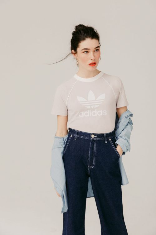 Model wearing a white Adidas t-shirt and dark denim trousers with a light denim shirt draped around arms against a neutral background.