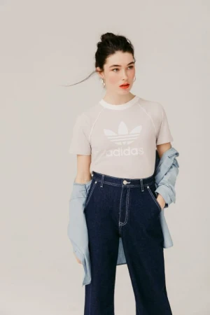 Model wearing a white Adidas t-shirt and dark denim trousers with a light denim shirt draped around arms against a neutral background.