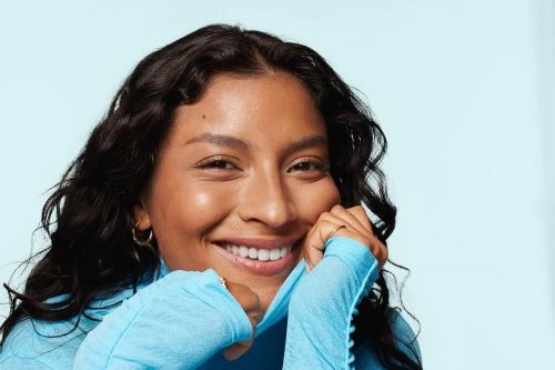Smiling young woman with tan skin and wavy black hair wears a blue long-sleeved shirt in a close-up studio portrait.