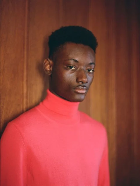 Portrait of a young Black man indoors with wooden paneling, wearing a red turtleneck sweater, gazing directly at the camera.