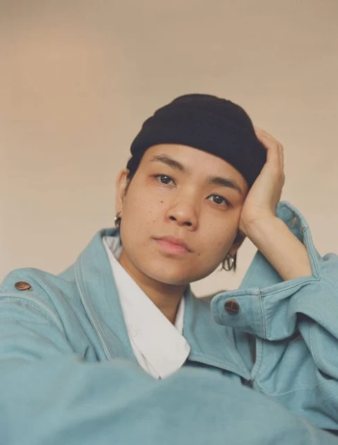 Headshot portrait of a young adult with a black beanie, wearing a white shirt and blue denim jacket, resting their head on their hand.