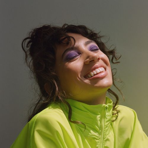 Close-up of a smiling adult woman with purple eyeshadow wearing a neon green jacket, looking upward in an indoor setting with natural lighting.