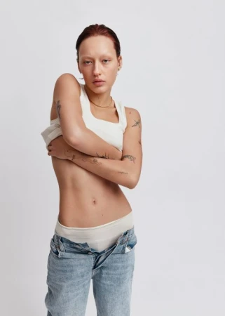 Model in white tank top pulling down unbuttoned blue denim jeans against a white seamless background