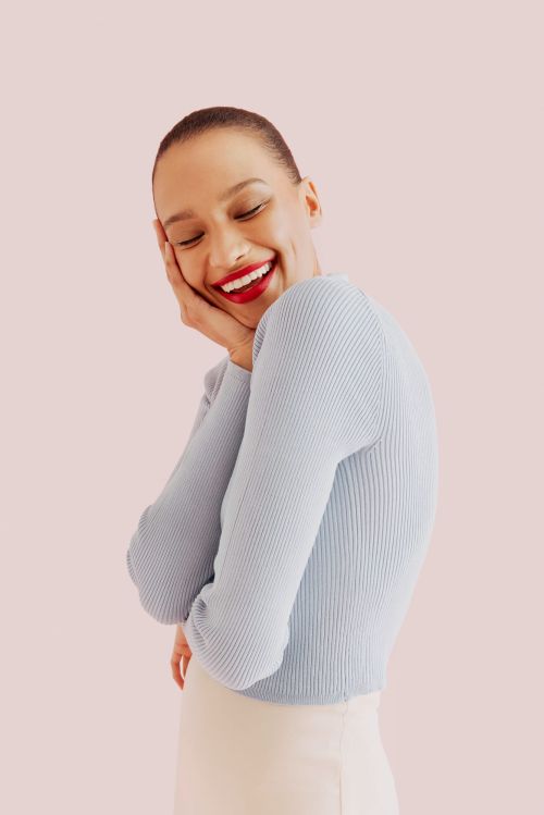 Smiling woman with hand on face wearing a light blue ribbed sweater and cream colored pants in a studio setting with soft lighting.