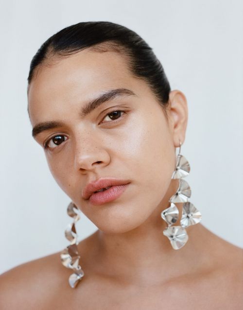 Close-up of a woman with dark hair and light skin wearing large silver earrings against a white background in a studio setting.