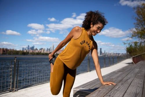 A woman in workout gear stretches her leg near a waterfront with a city skyline in the distance, under a bright blue sky.