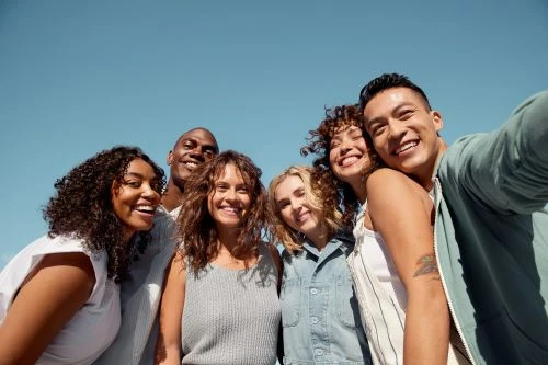 Six smiling diverse adults posing together in a selfie against a clear blue sky, captured with a wide-angle lens on a sunny day.