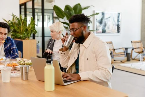 A man drinks from a metal cup while working on a laptop at a table with two other people at a casual business lunch.
