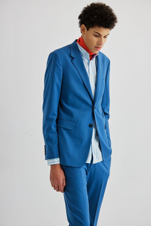 Young adult posing in a blue suit with an open blue and white striped shirt and a red scarf against a white backdrop in a studio.