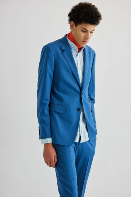 Young adult posing in a blue suit with an open blue and white striped shirt and a red scarf against a white backdrop in a studio.