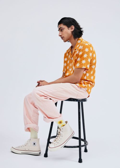 A young adult male sits on a black stool in a studio, wearing a patterned orange shirt, pink pants, and high-top sneakers.