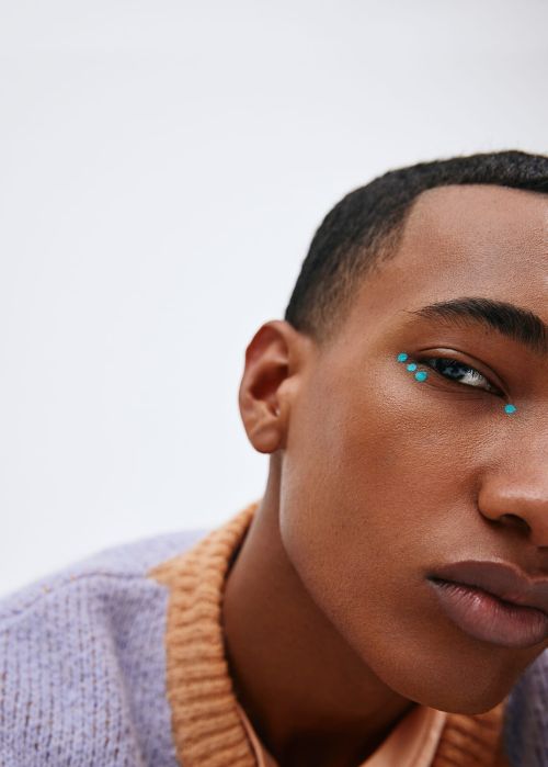 Close-up studio portrait of a young adult male of African descent with minimalist turquoise makeup around the eye against a white backdrop.