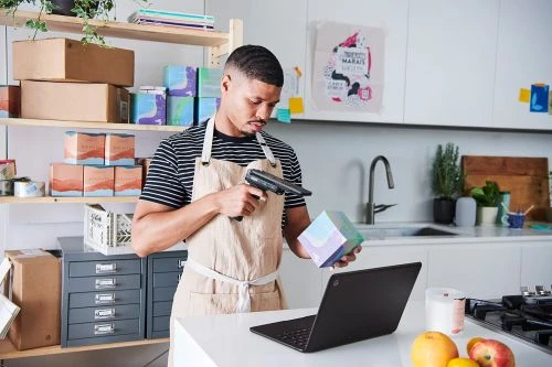 Man in apron scans cube with scanner gun in kitchen, laptop on counter with fruit, packages stacked on shelves in background.