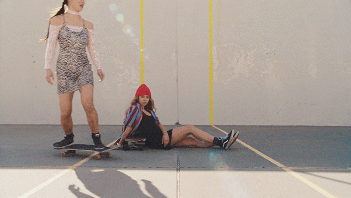 Two young women in casual summer outfits pose with a skateboard against a gray concrete wall with yellow lines. Youth lifestyle theme.