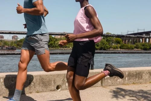 Two athletes run side-by-side along a waterfront path on a sunny day. A river and cityscape with a bridge are in the background.