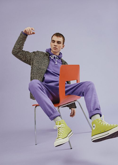 A young adult sits on an orange chair in a purple hoodie and sweatpants, lime green sneakers, and a leopard print jacket, on a purple background.