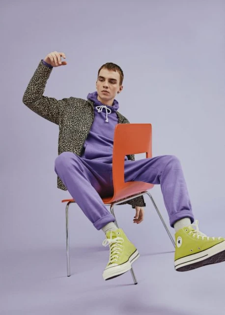 A young adult sits on an orange chair in a purple hoodie and sweatpants, lime green sneakers, and a leopard print jacket, on a purple background.